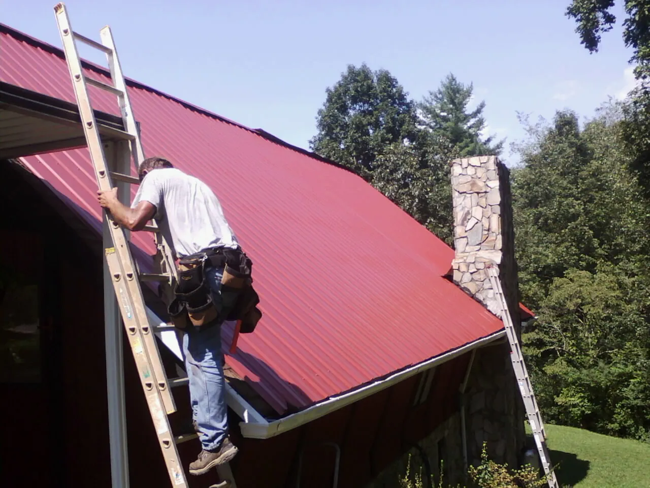 Roofer on a ladder beside a red metal roof and stone chimney — Soffit & Fascia Repair by Roofing Brothers in Minden City